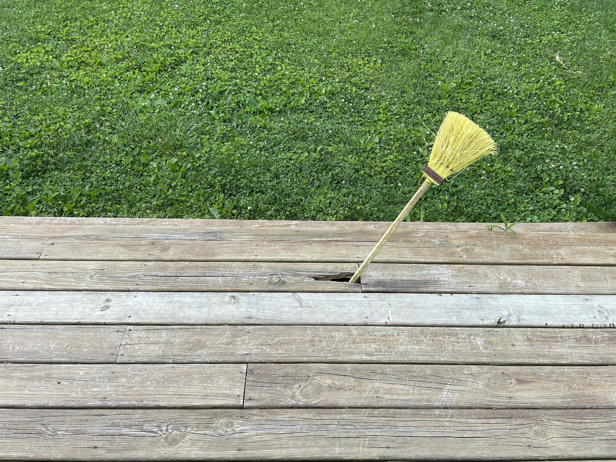 A yellow broom sticking out of a hole in a wooden deck with green grass behind it.