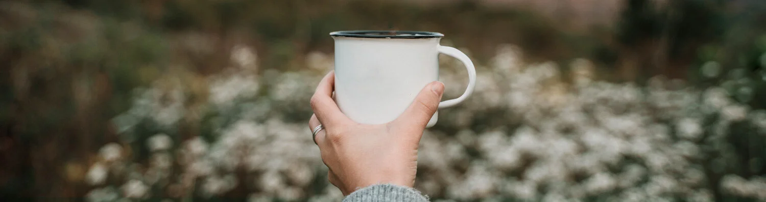 A person holding up a white tea mug against a background of fall foliage