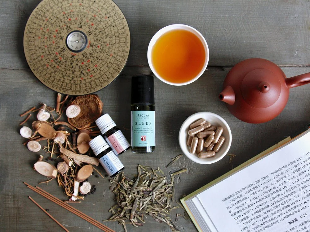 Traditional Chinese herbs, essential oil blends, and a yixing teapot next to an open book on Chinese Medicine