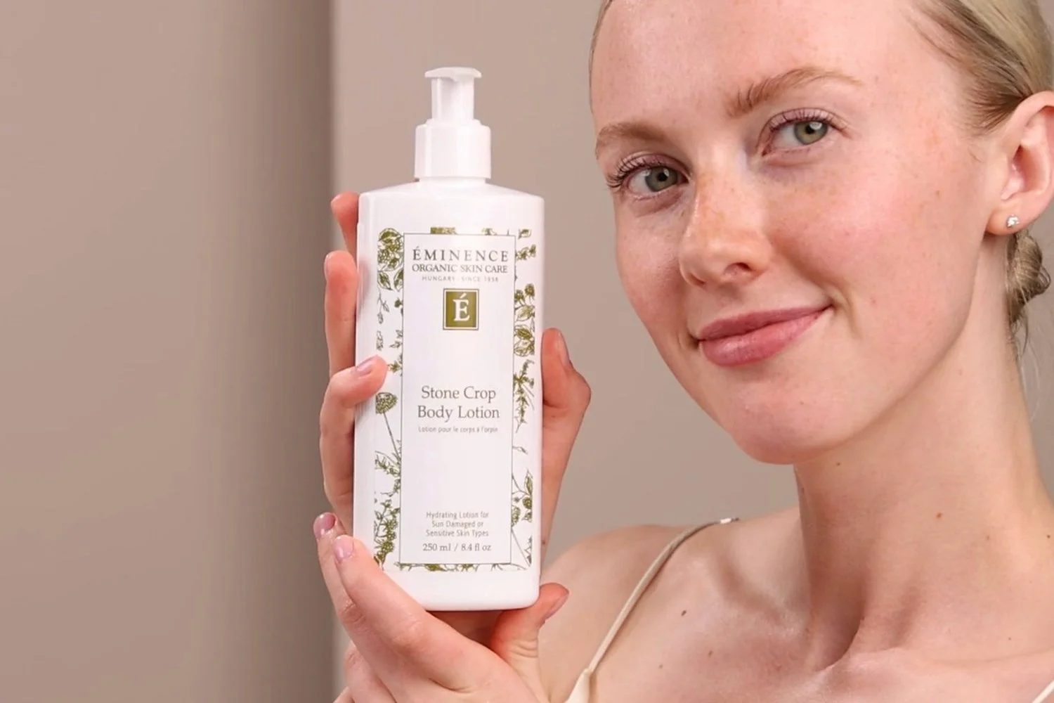 A woman holding the Eminence Organics Stone Crop Body Lotion bottle next to her face
