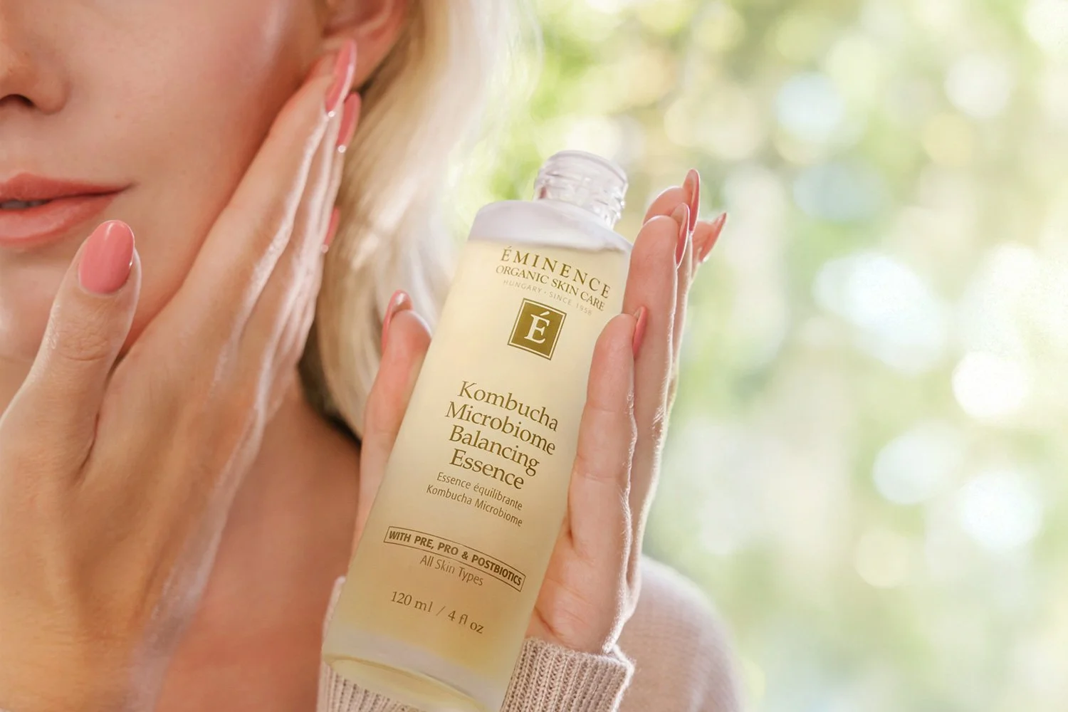 A woman holding the Eminence Organics Kombucha Microbiome Balancing Essence glass bottle and applying the essence onto her face