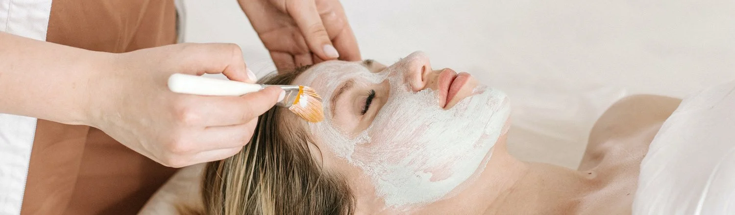 Esthetician using a soft brush to apply a mask to a woman’s face during a facial