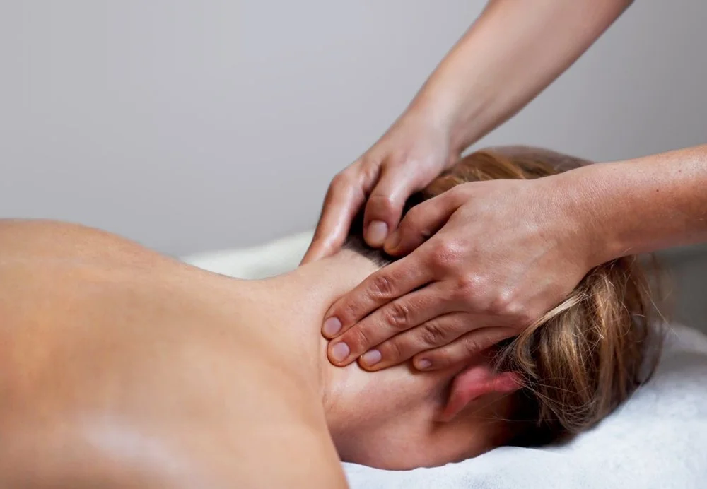 A woman receiving a neck massage. The massage therapist uses both hands to apply pressure on muscles near the base of the skull