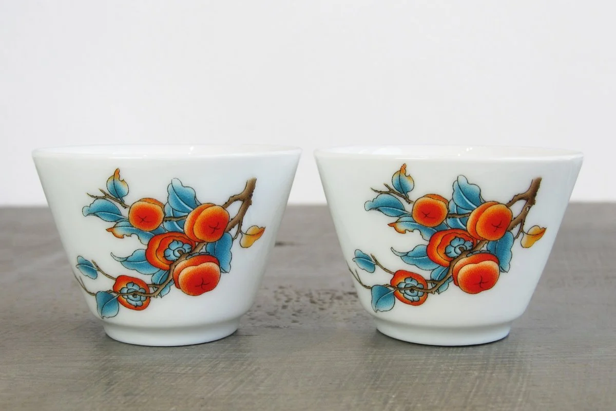 Close-up of two white glaze teacups decorated with a vibrant persimmon branch motif