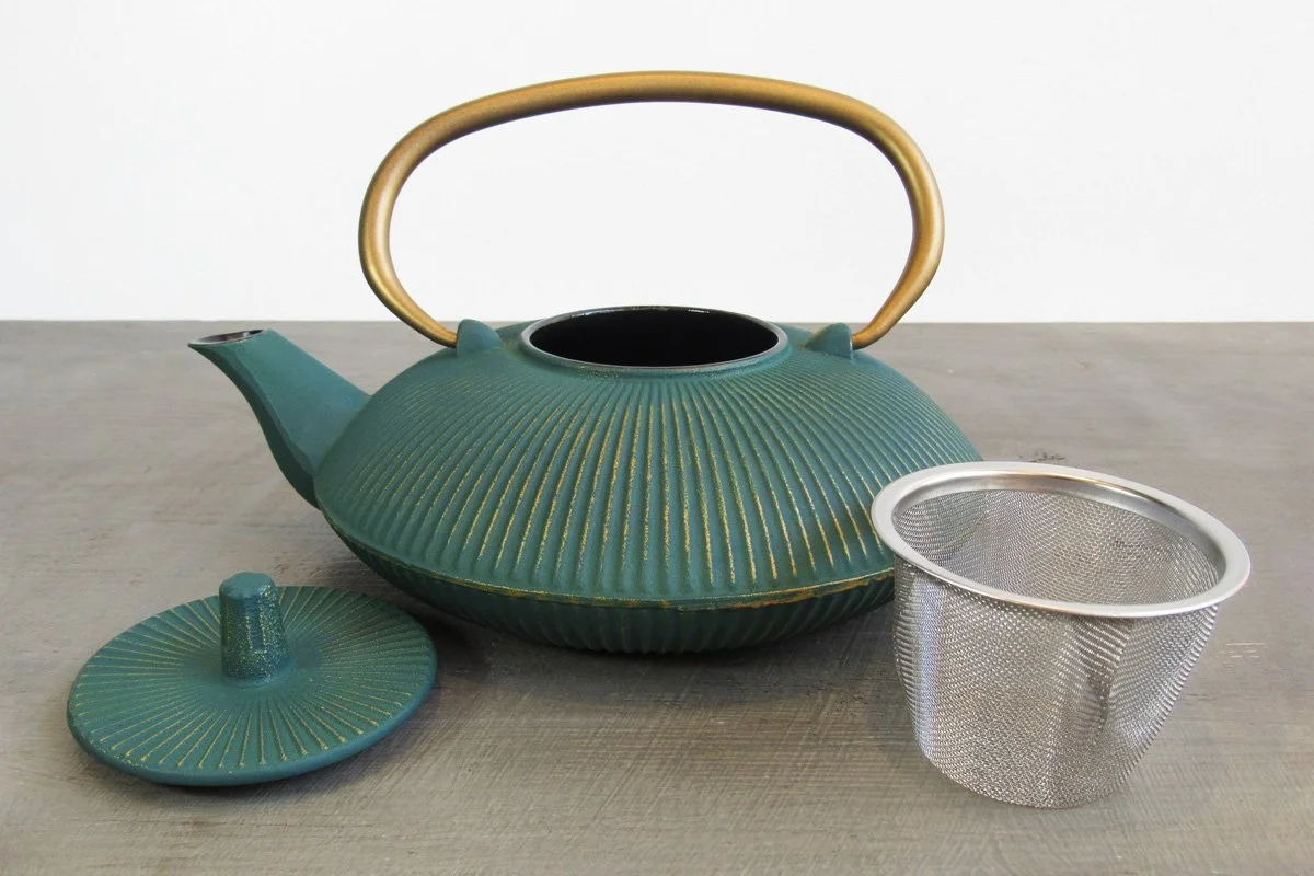 Open teal cast iron teapot with a gold handle next to its lid and stainless steel infuser basket