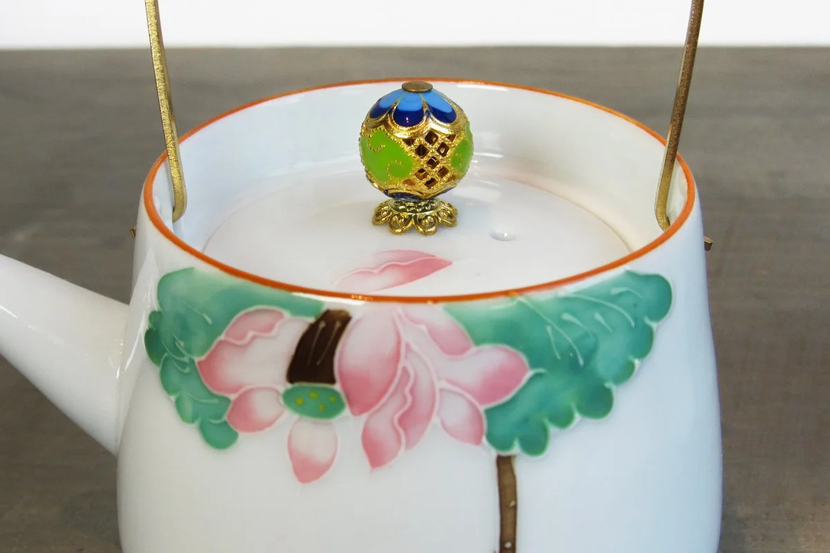Close-up of the lid of a white teapot with an ornate gold knob and hand-painted lotus flower design