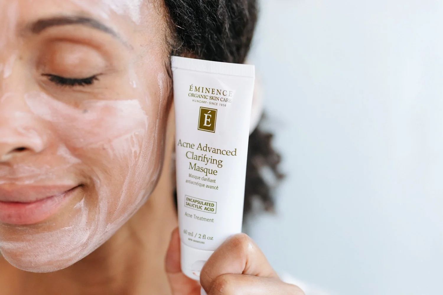 A woman holding the Acne Advanced Clarifying Masque container next to her face while the mask dries on her skin