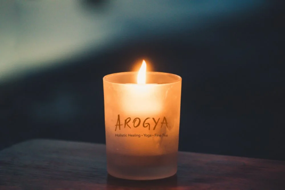 Lit candle in a dark room branded with the Arogya Holistic Healing logo