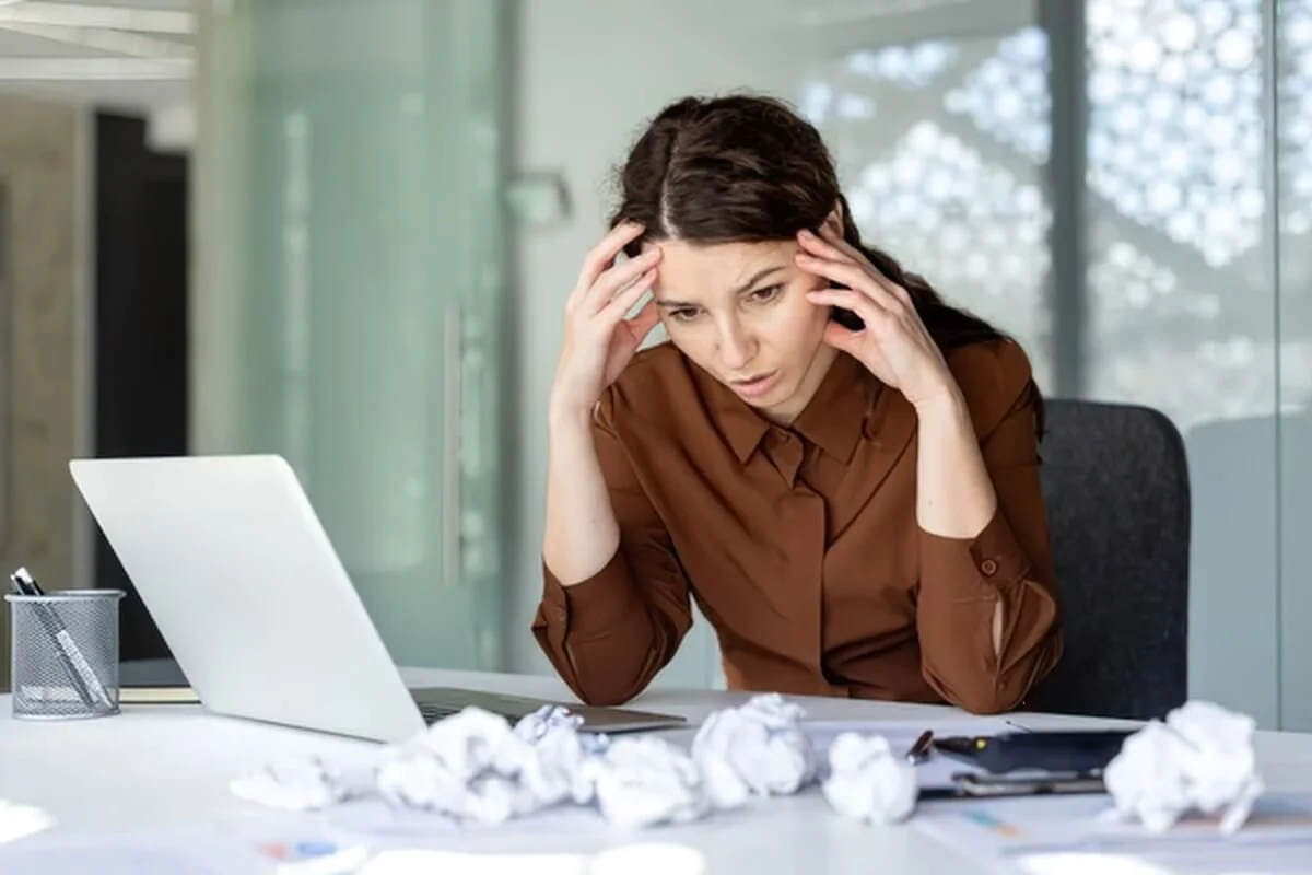 Overwhelmed woman at a desk of crumpled papers, hands on head, a common reason adults seek an ADHD evaluation
