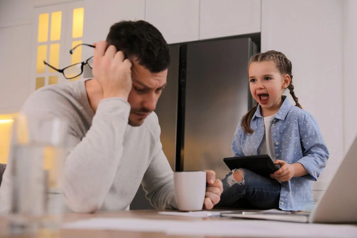 Frustrated man holding glasses to his head while drinking coffee at his laptop as a young girl screams beside him representing the overwhelm experienced by AuDHD families seeking ADHD and Autism evaluations at True Reflections MHS in Middlesex NJ