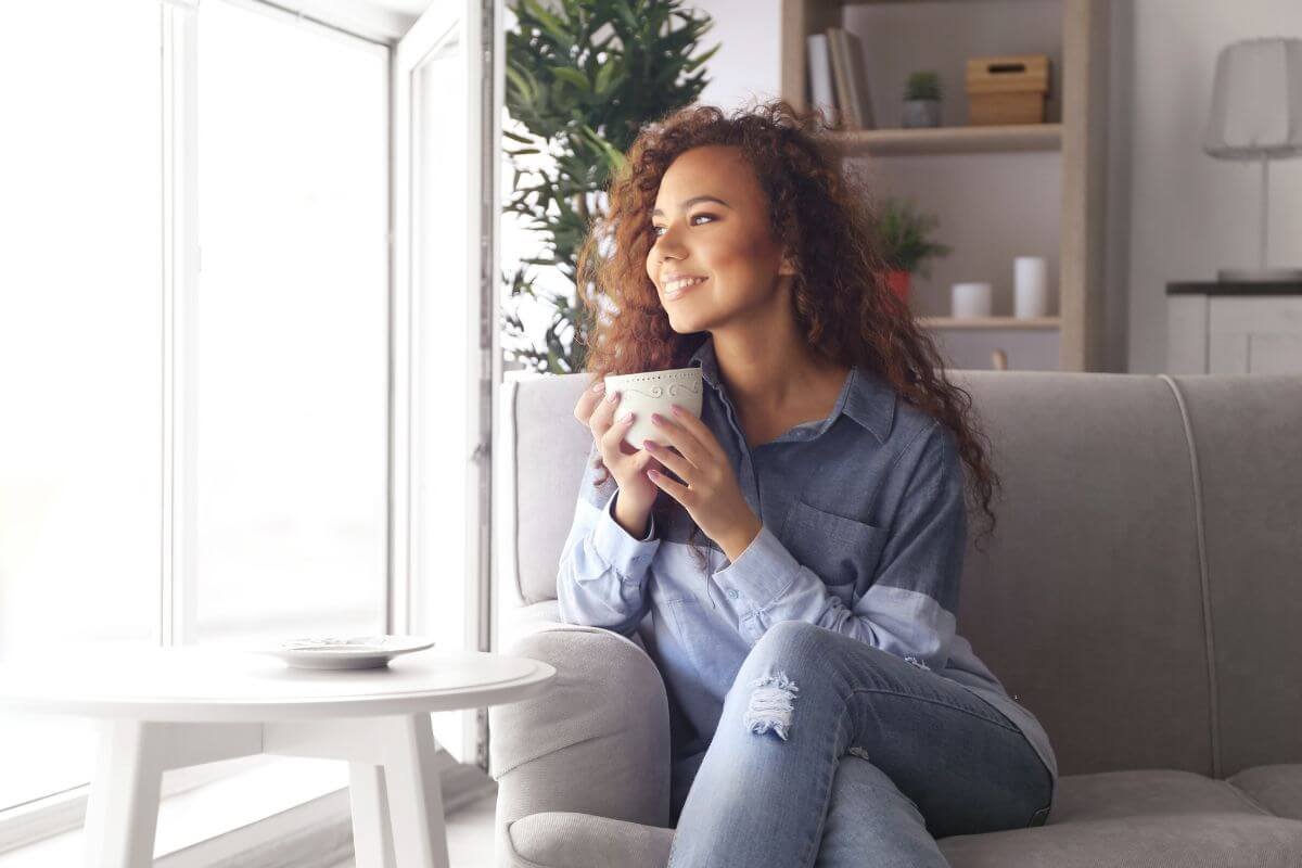 A woman sitting on a couch smiling and drinking coffee while looking out a window representing the calm and relief that can follow an ADHD or autism diagnosis as an adult in New Jersey