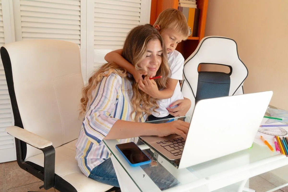 Mother working on laptop while young son hugs her and holds a colored pencil, representing a parent researching ADHD testing for her child in Middlesex, NJ