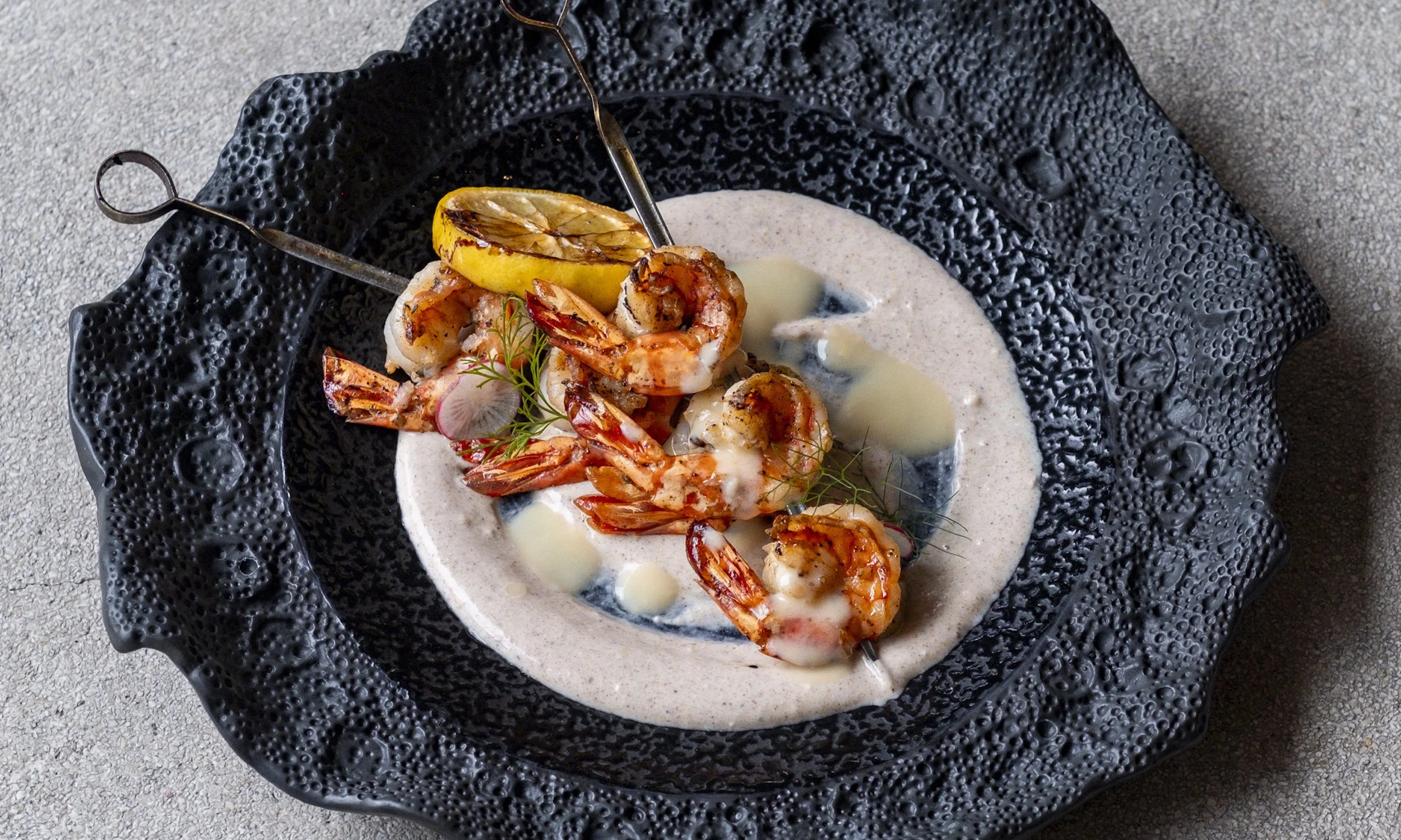 Seafood dish with grilled shrimp and lemon on a black textured plate with creamy sauce and garnishes.