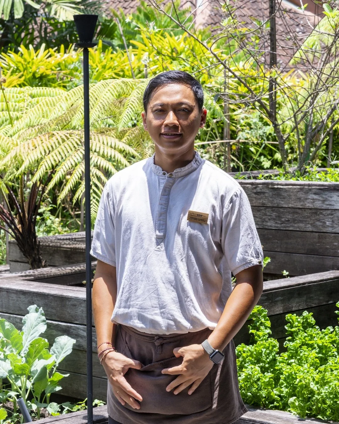 A man standing outdoors in a garden with lush green plants, wearing a white chef's jacket with a name tag, and a smartwatch on his left wrist.