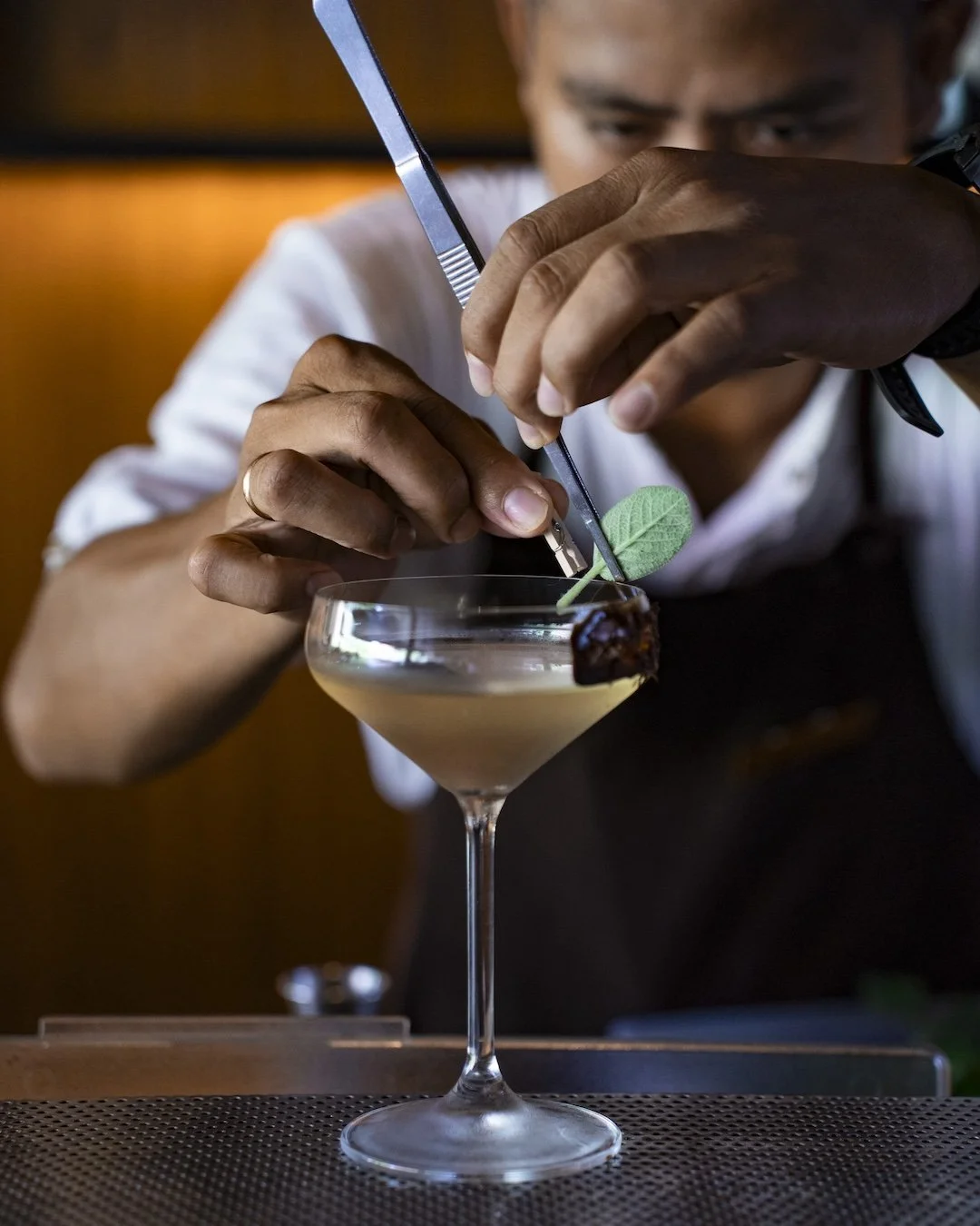 A bartender garnishing a cocktail with a green leaf using tweezers in a coupe glass.