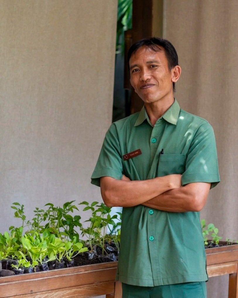A man with short dark hair wearing a green uniform with a name tag, standing with arms crossed next to a wooden table with potted plants. He is smiling slightly and looking at the camera.