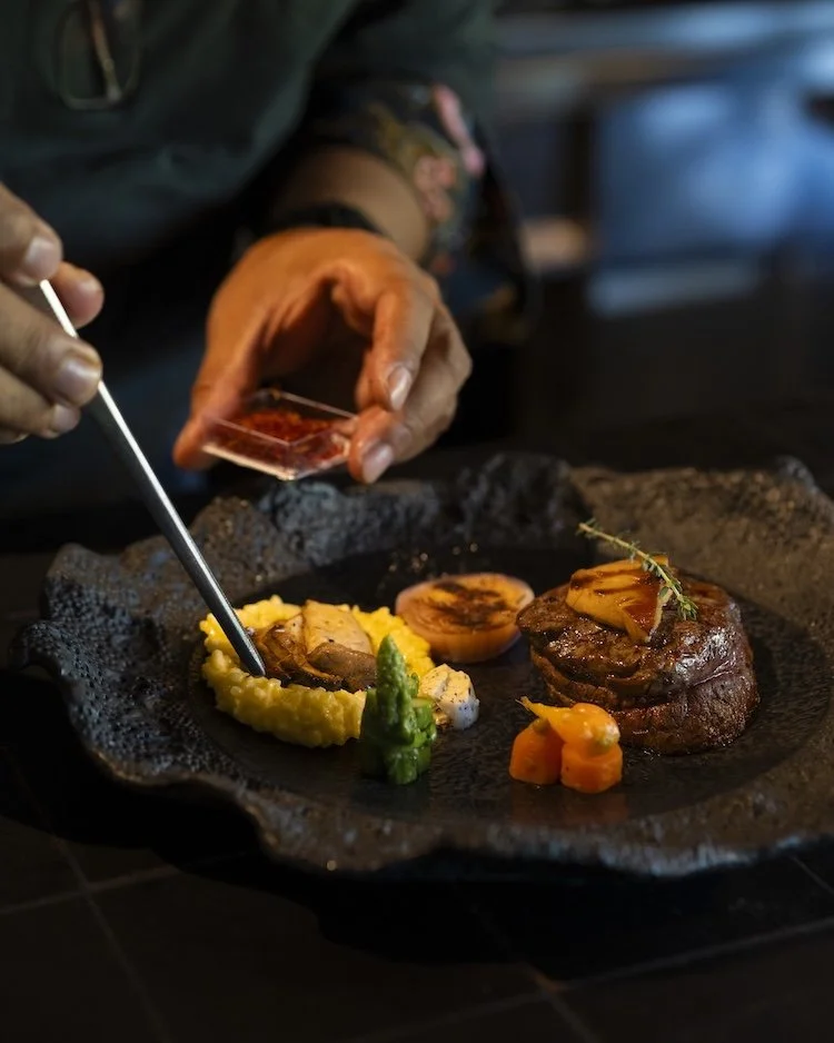 A chef preparing a plated steak dish with mashed potatoes, grilled tomato, and vegetables in a restaurant kitchen.