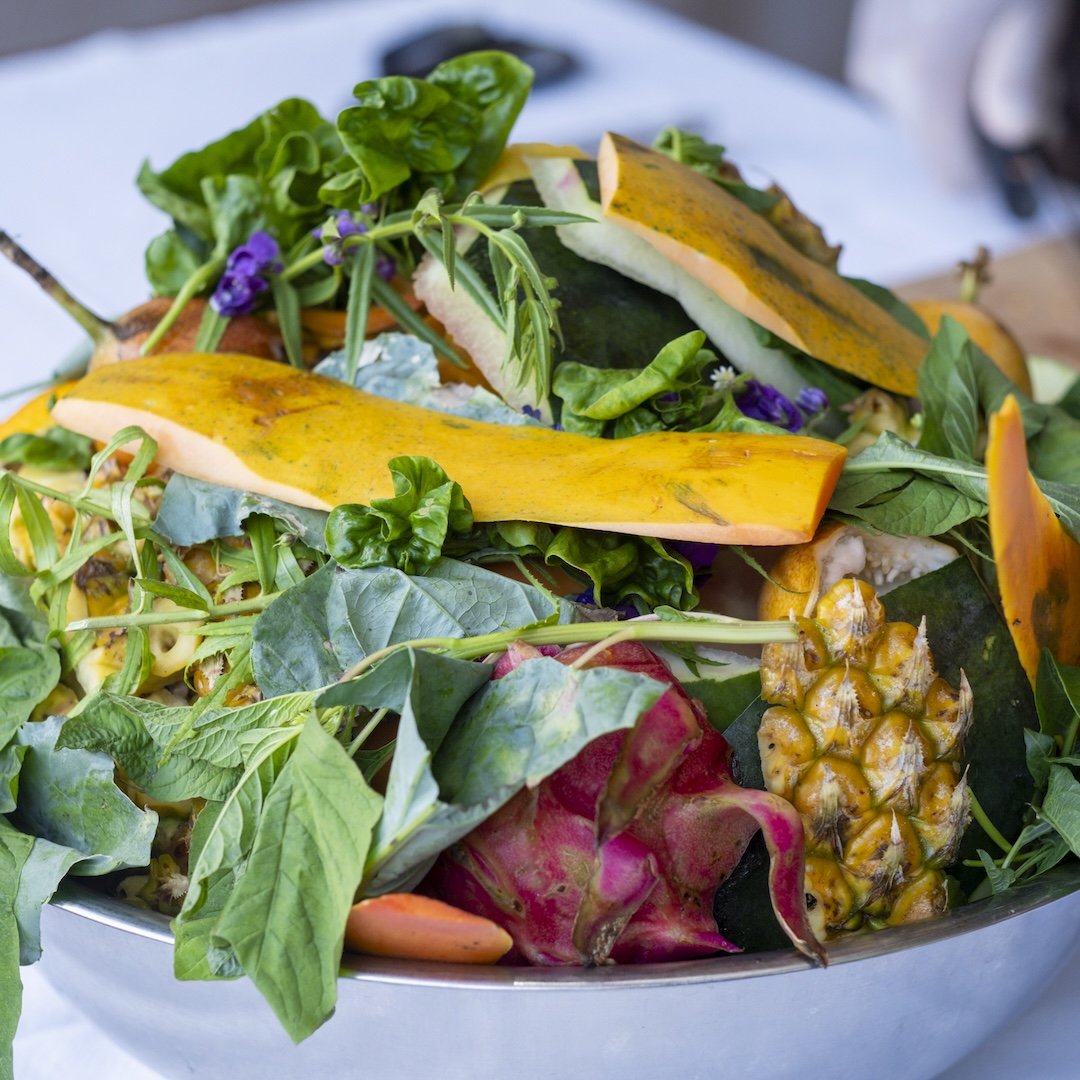 Fresh tropical fruits including pumpkin slices, pineapple, dragon fruit, and leafy greens in a metal bowl.