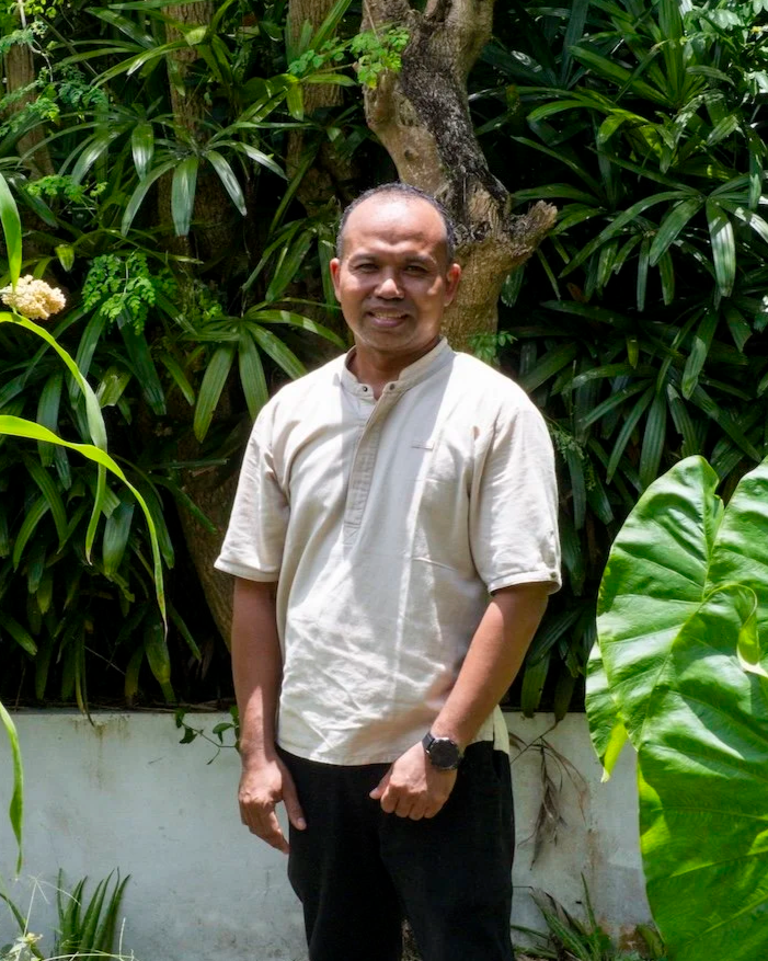 A man standing outdoors surrounded by lush green plants and trees, wearing a beige shirt and black pants, smiling at the camera.