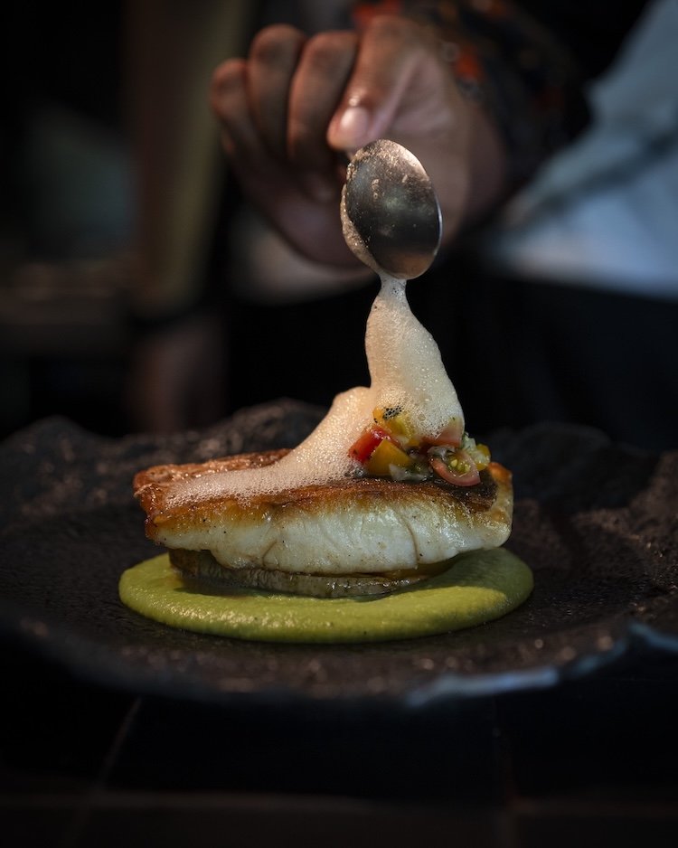 Close-up of a chef pouring foam over a plated fish dish with vegetables, with green sauce underneath on a black plate.