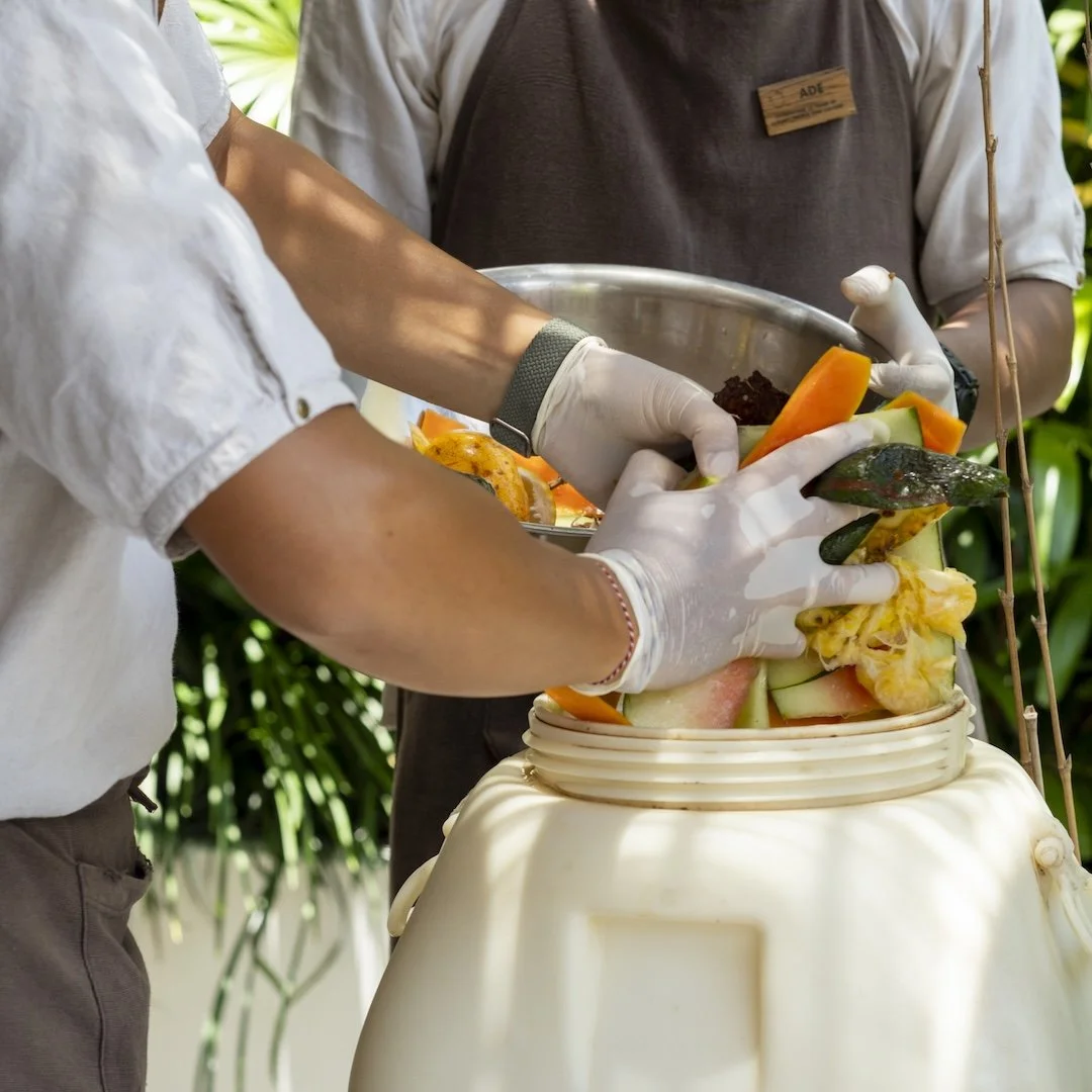 Two people wearing gloves preparing fruit salad with various cut fruits in a bowl, in an outdoor setting.