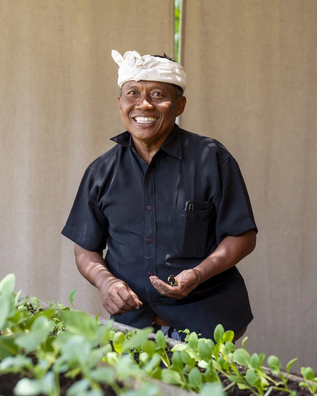 A smiling man wearing a black shirt and a white headwrap, standing behind a tray of green seedlings.