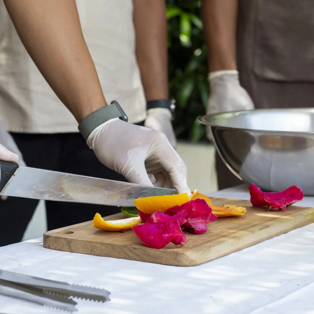 Person slicing colorful bell peppers on a wooden cutting board, wearing gloves, with a metal mixing bowl nearby.