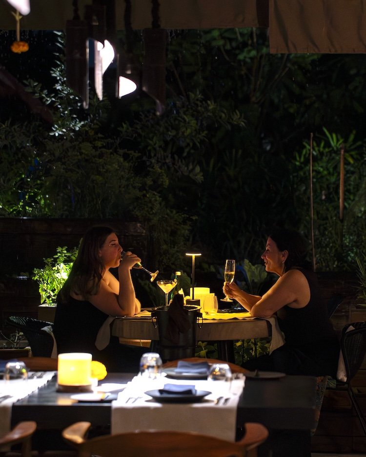Two people enjoying a candlelit dinner at an outdoor restaurant, with dim lighting and greenery in the background, both holding glasses and engaged in conversation.