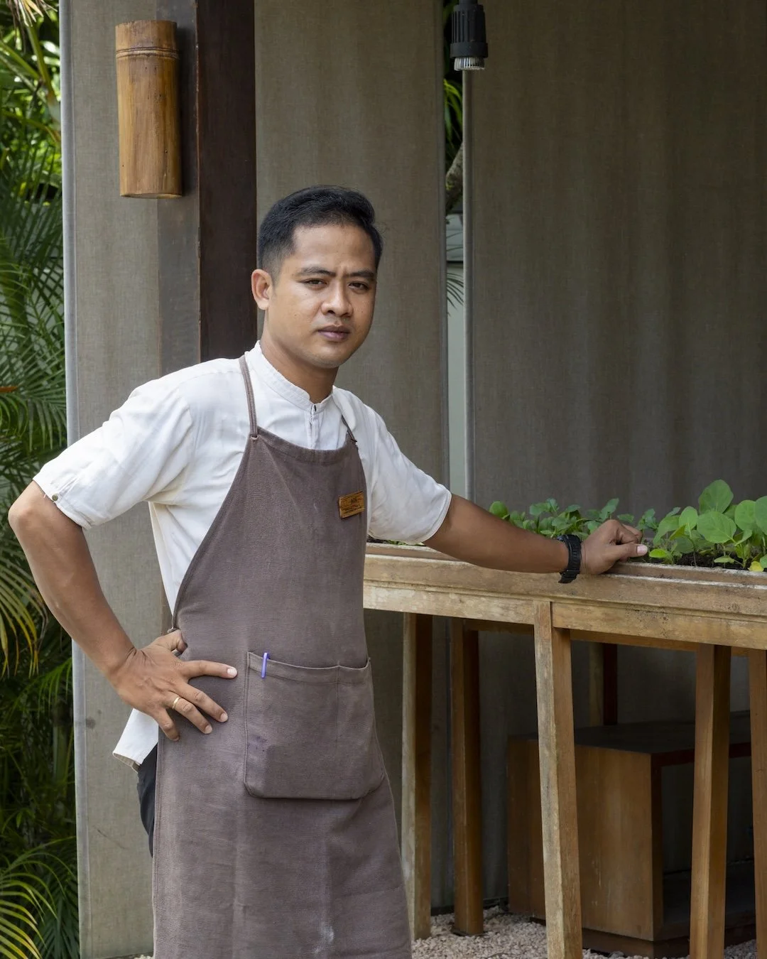 A man wearing a white shirt and a brown apron standing outdoors next to a wooden table with green plants. He has short black hair and a serious expression.