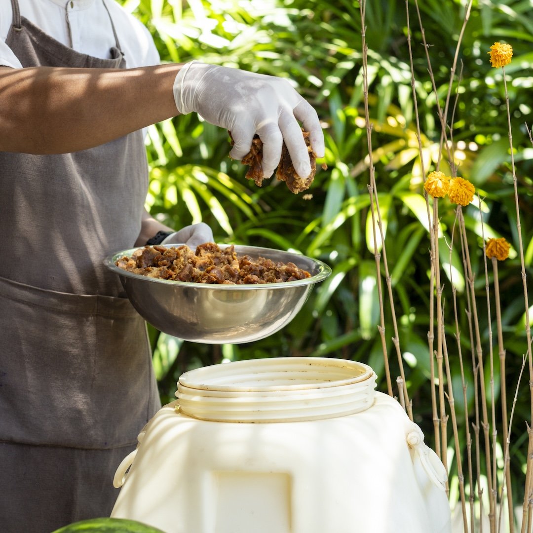Person wearing gloves scooping cooked meat into a bowl outdoors, surrounded by green plants and yellow flowers.