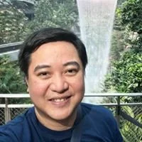 A woman smiling outdoors with lush greenery and a waterfall in the background.