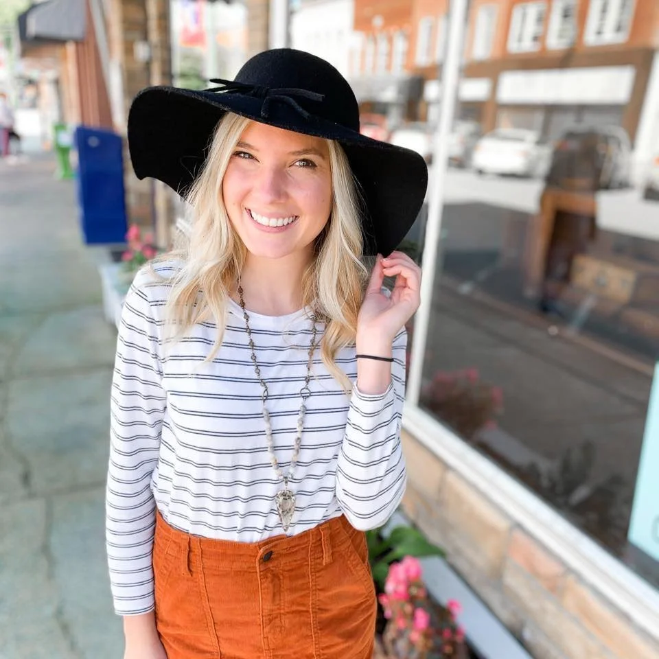 A woman with blonde hair wearing a large black sunhat, a white and black striped long-sleeve shirt, orange pants, and a necklace with a pendant, standing outside near a storefront with a large window.