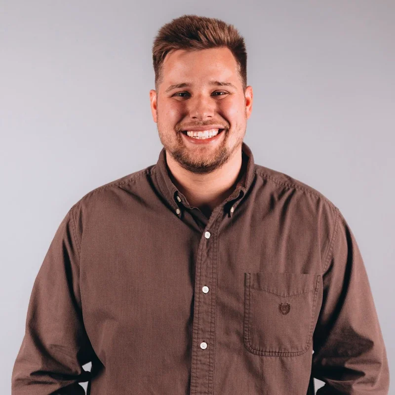 A young man with short brown hair and a goatee, smiling, wearing a brown button-up shirt against a light gray background.