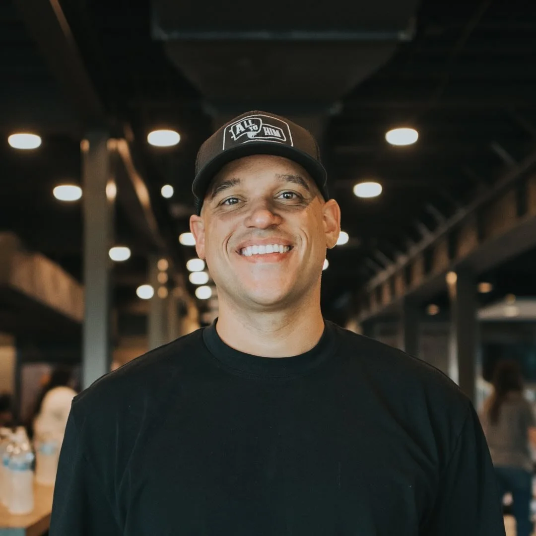 Smiling man wearing a black T-shirt and a black cap in a modern indoor setting with ceiling lights and blurred background with people.
