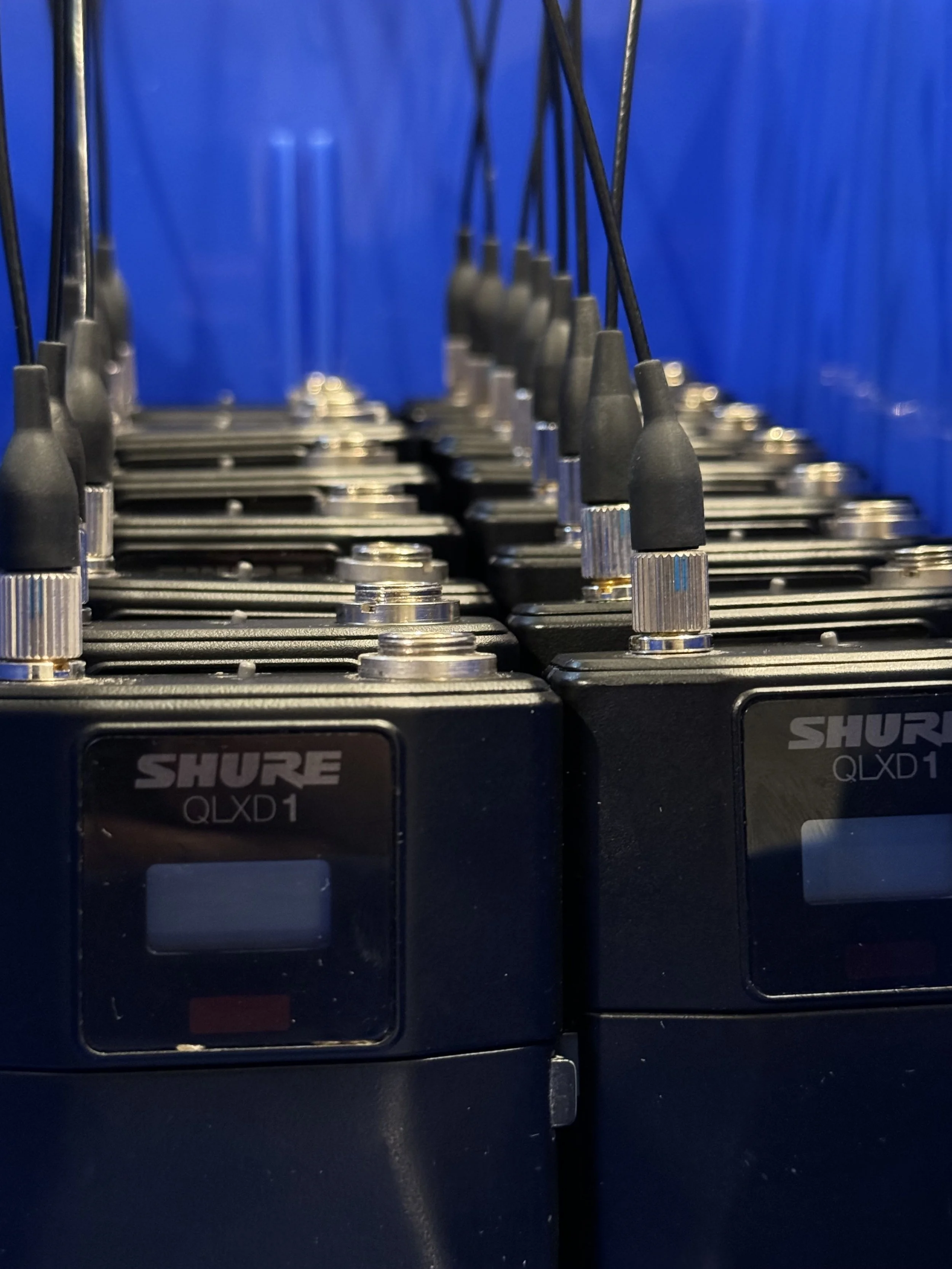Close-up view of multiple Shure QLXD1 wireless microphone transmitters, arranged in a row, with blue background.