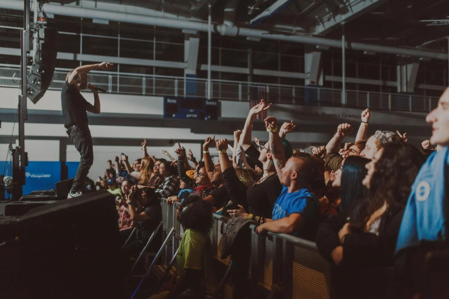 A crowd of concert attendees raising their hands while watching a performer on stage inside a large indoor venue with industrial ceiling and railings.