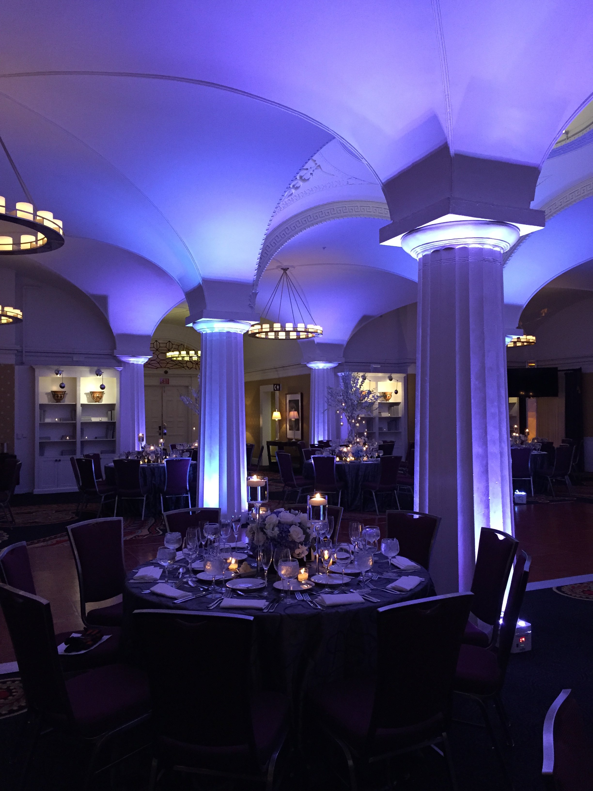 Elegant banquet hall decorated for an event with white columns lit by blue lighting, round tables with centerpieces, candles, and elegant place settings, and a high ceiling with ornate molding and chandeliers.