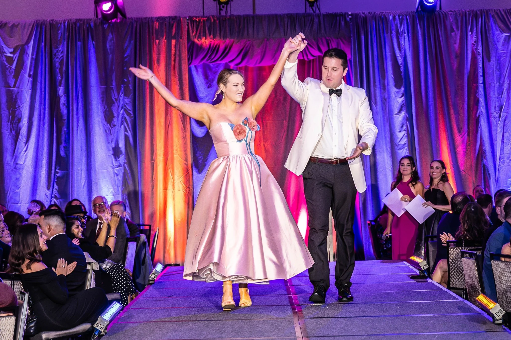 A man and woman in formal attire walking down a runway, holding hands with hands raised. The woman wears a pink strapless dress with floral accents, and the man wears a white jacket, white shirt, and black bow tie. They are smiling, surrounded by an audience and colorful stage curtains.