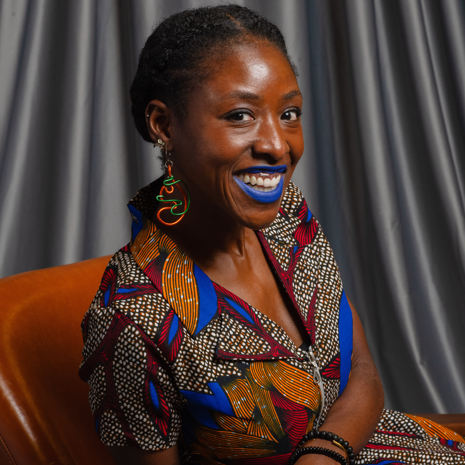 Black woman sitting in a chair looking at the camera, smiling, wearing blue lipstick