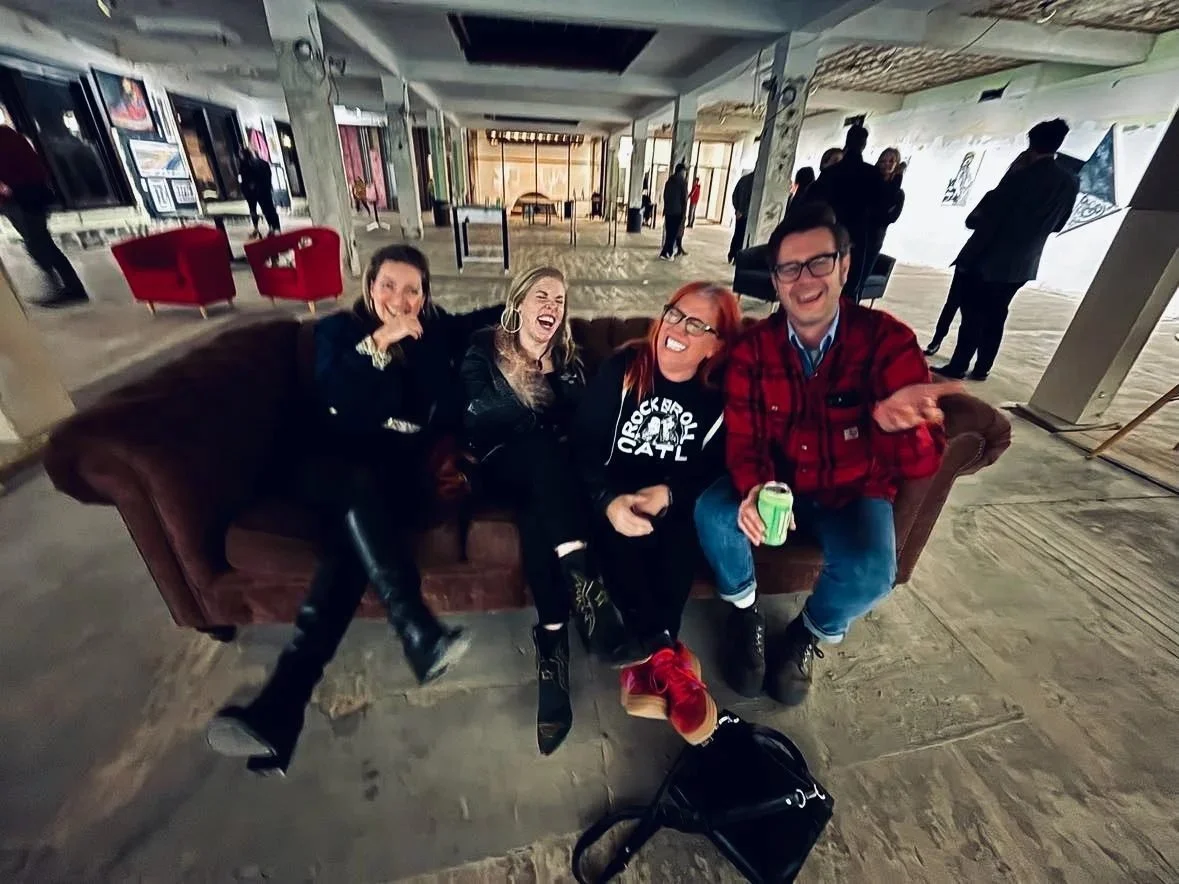 Four people laughing on a couch in the middle of an empty downtown floor hung with art.