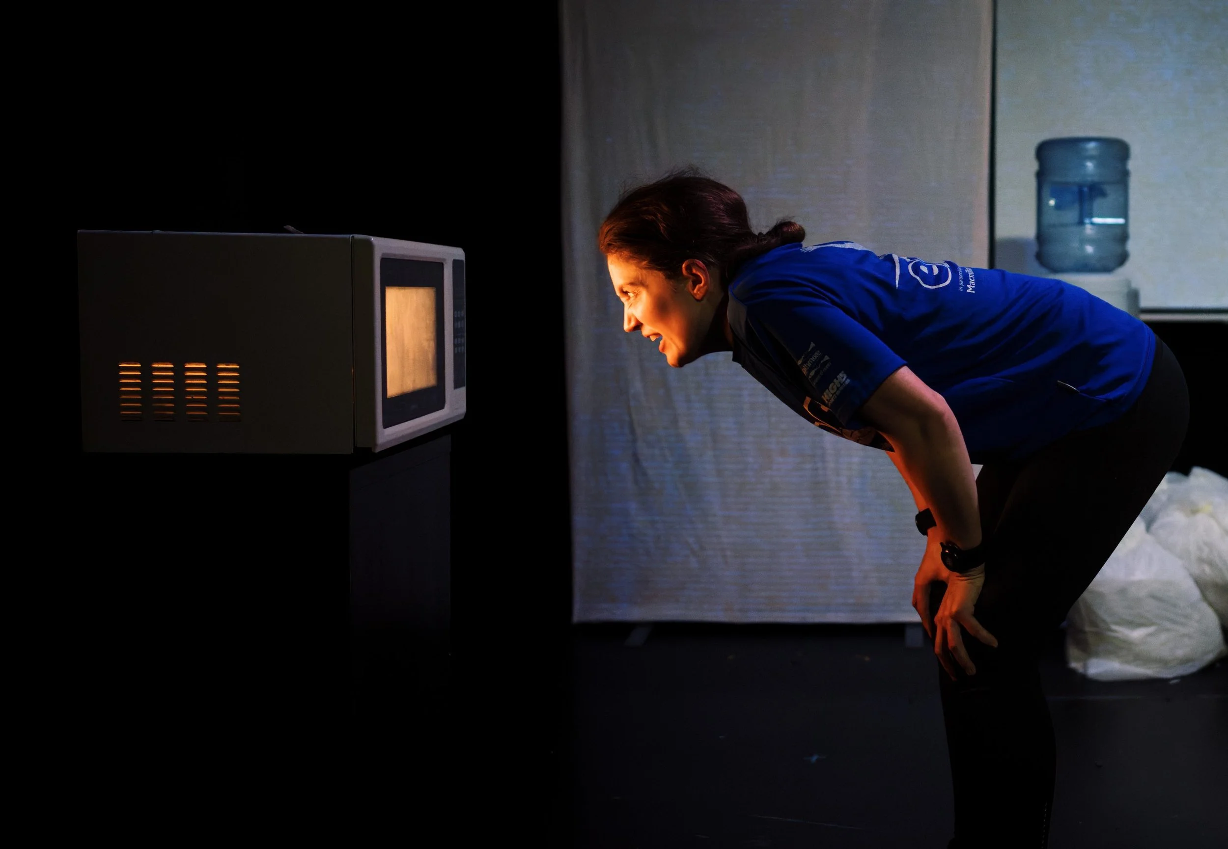 a woman wearing a blue tshirt stands leaning down to look in the door of a microwave. the microwave is lit up inside. The woman’s face is lit up with the warm light from the microwave.