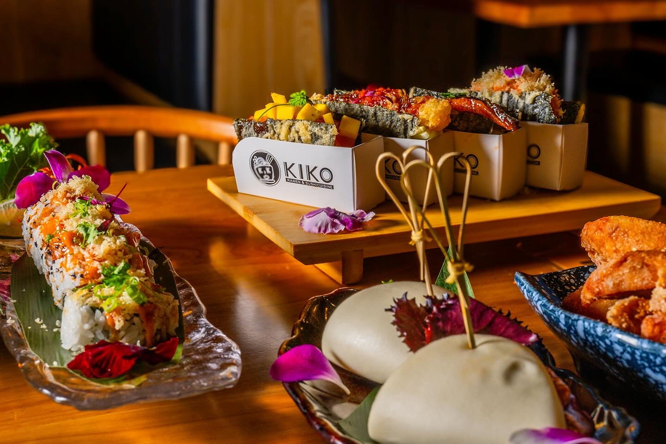 Assorted sushi rolls, tempura, and fried chicken on a wooden table with decorative flowers and leaves.