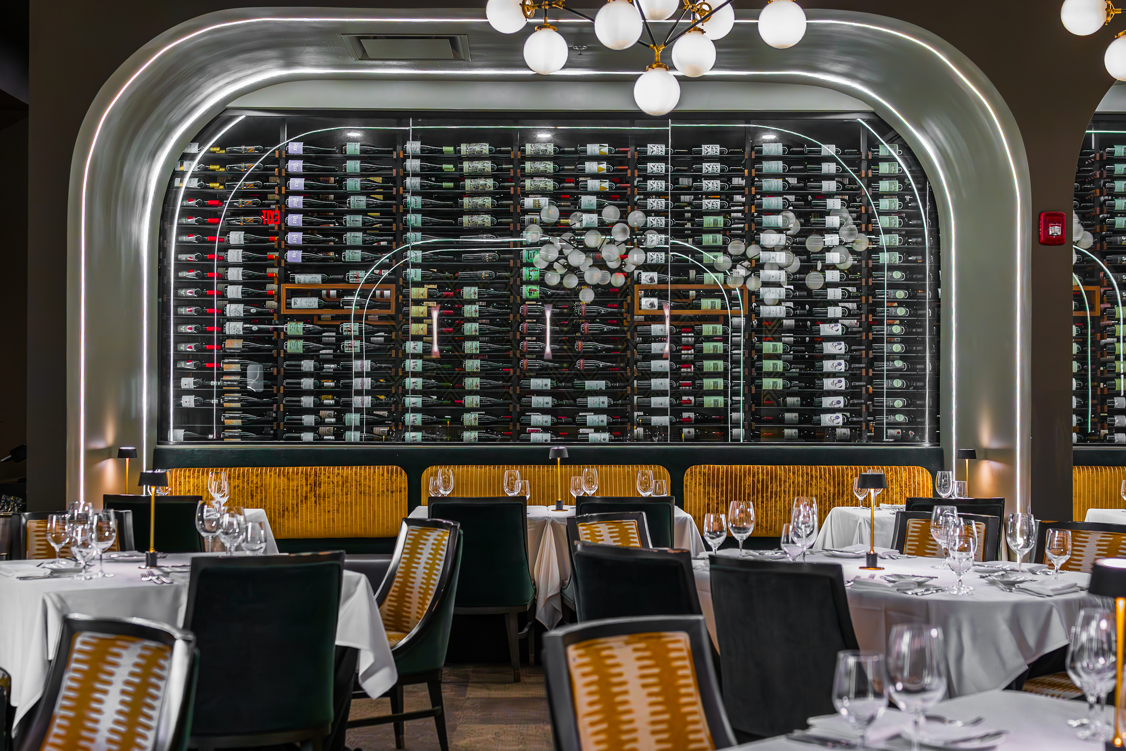 Elegant restaurant interior with white-draped tables set with wine glasses and cutlery, a stylish black and gold chandelier, and a large wine display behind glass with bottles organized on shelves.