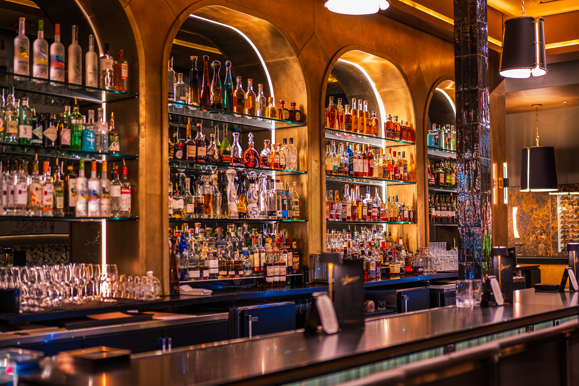 A well-stocked bar with various bottles of liquor displayed on illuminated glass shelves behind the counter, in a upscale setting with warm lighting and decorative elements.