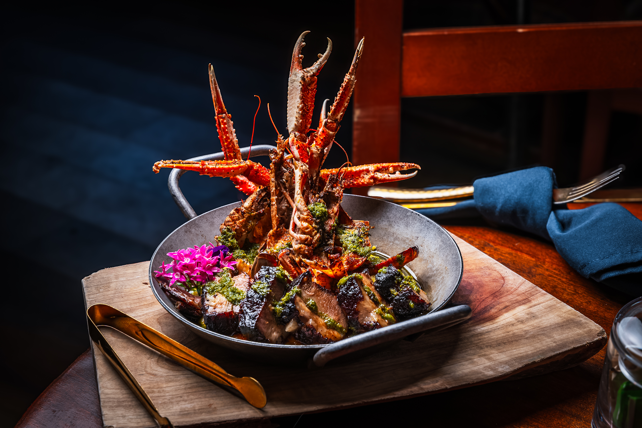 A seafood dish featuring lobster claws and lobster tails garnished with green herbs and pink flowers, served in a metal skillet on a wooden board, with gold-colored cutlery and a dark background.