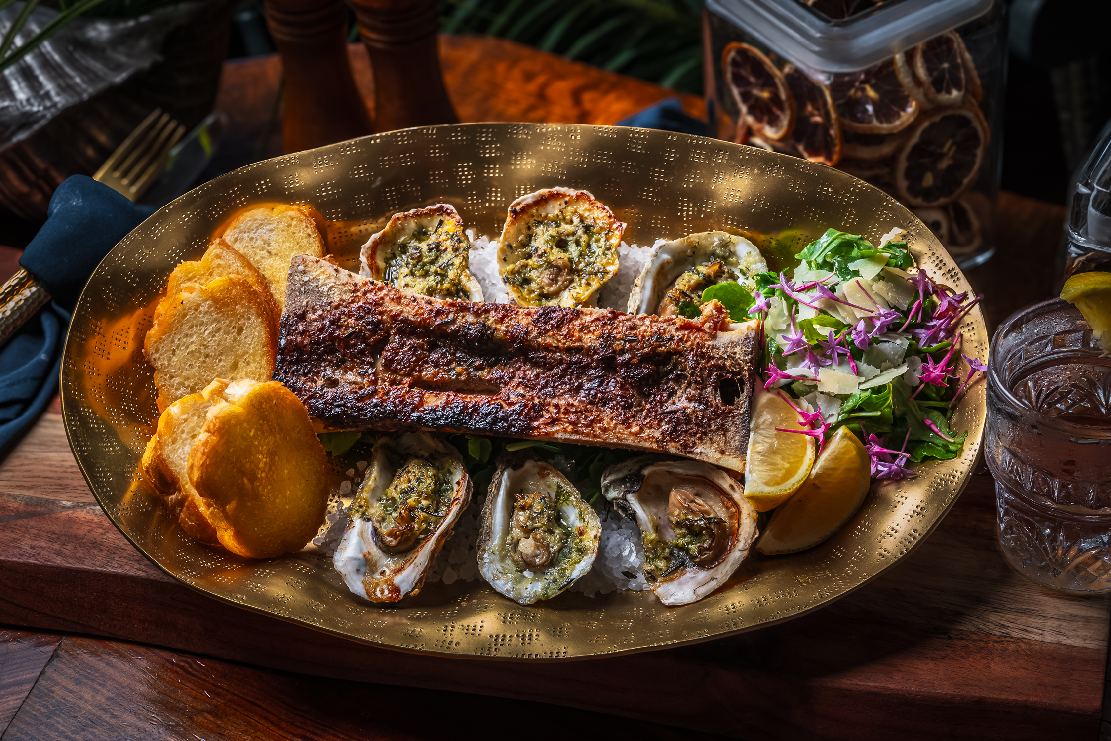 A seafood platter featuring grilled fish, oysters on ice, garlic bread, lemon wedges, and a side salad with purple flowers.