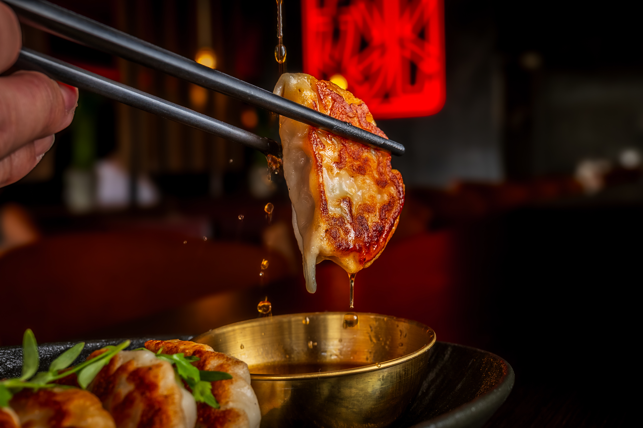 Close-up of a hand using chopsticks to pick up a crispy, golden-brown gyoza with melted cheese and sauce dripping from it, served on a black plate with a small metallic bowl of dipping sauce.