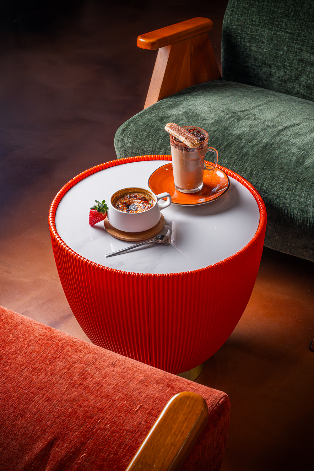 A small round red side table with a white surface holds a mug of baked-smort crème brûlée with a strawberry garnish and a tall glass of hot chocolate topped with churro sticks. The table is in front of a green armchair and a red upholstered sofa.