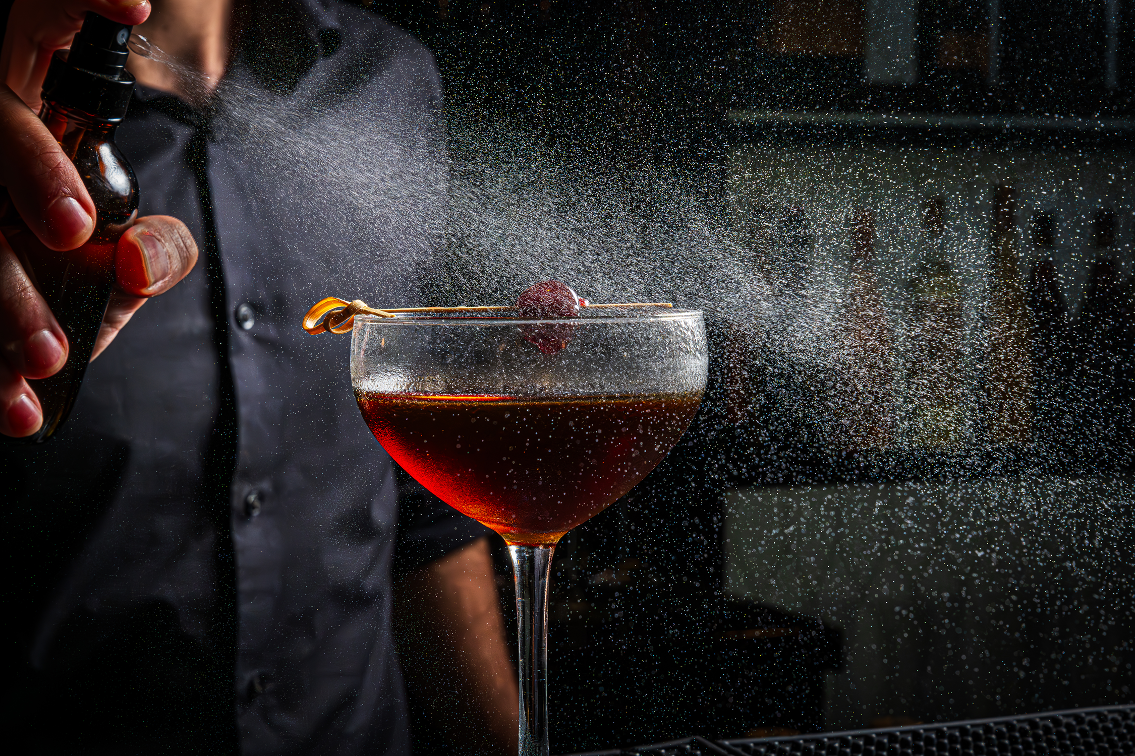 A bartender spraying a cocktail with a spray bottle over a glass of red drink garnished with a cherry on a skewer, with a fine mist of liquid around.
