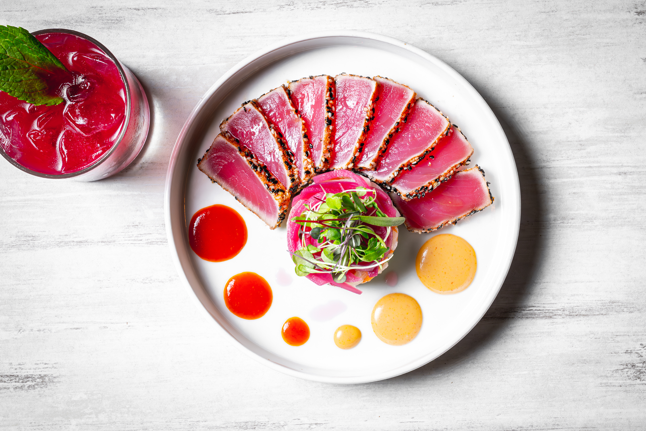 Seared tuna sashimi served on a white plate with microgreens, pink pickled vegetables, and with several dollops of yellow and red sauces, alongside a glass of red beverage with mint.